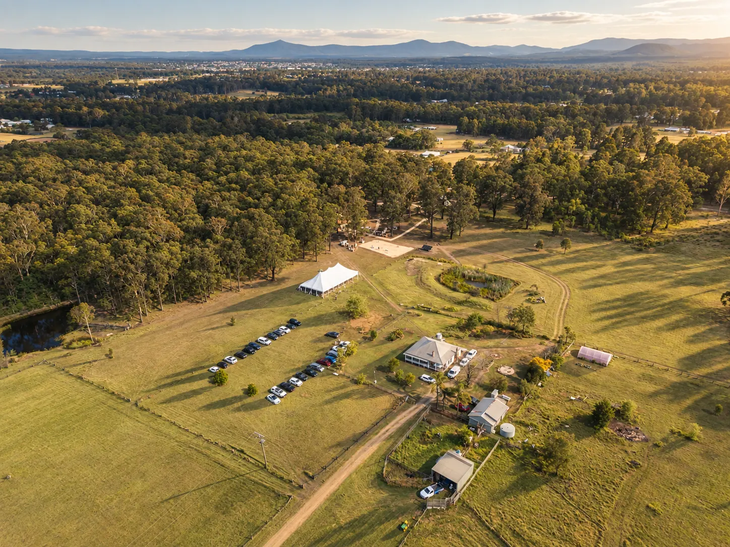 Aerial view of a house, white wedding marquee, parked cars and open paddocks surrounded by trees.
