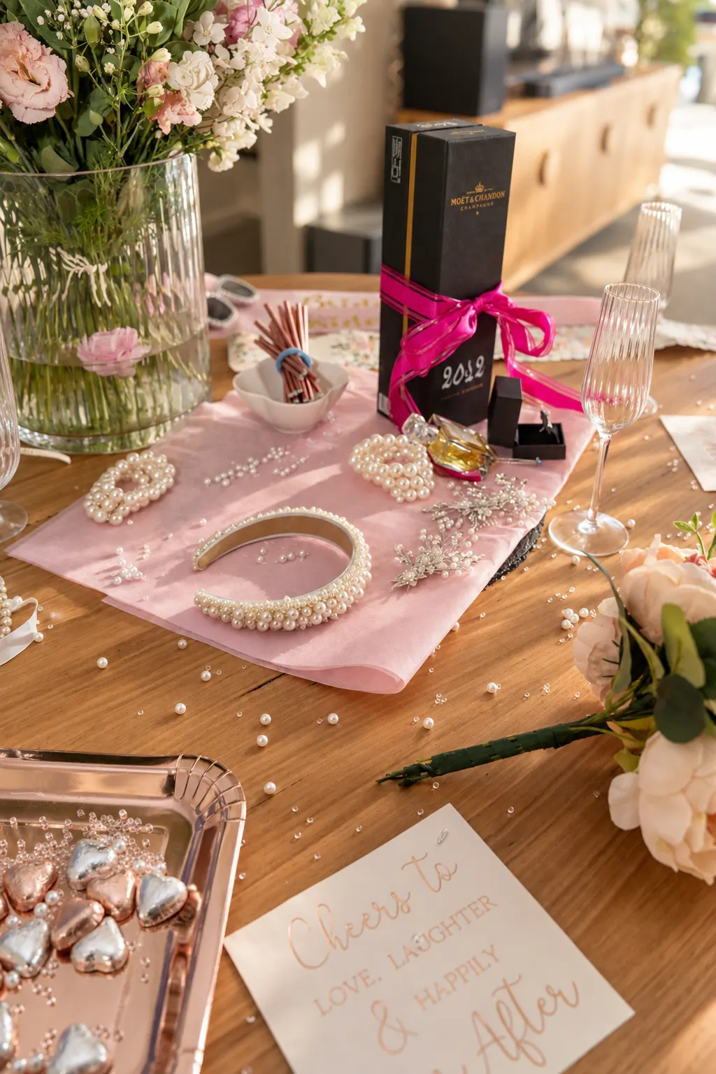 Pearl bridal accessories, flowers, a boxed champagne bottle and glasses arranged on a wooden table.