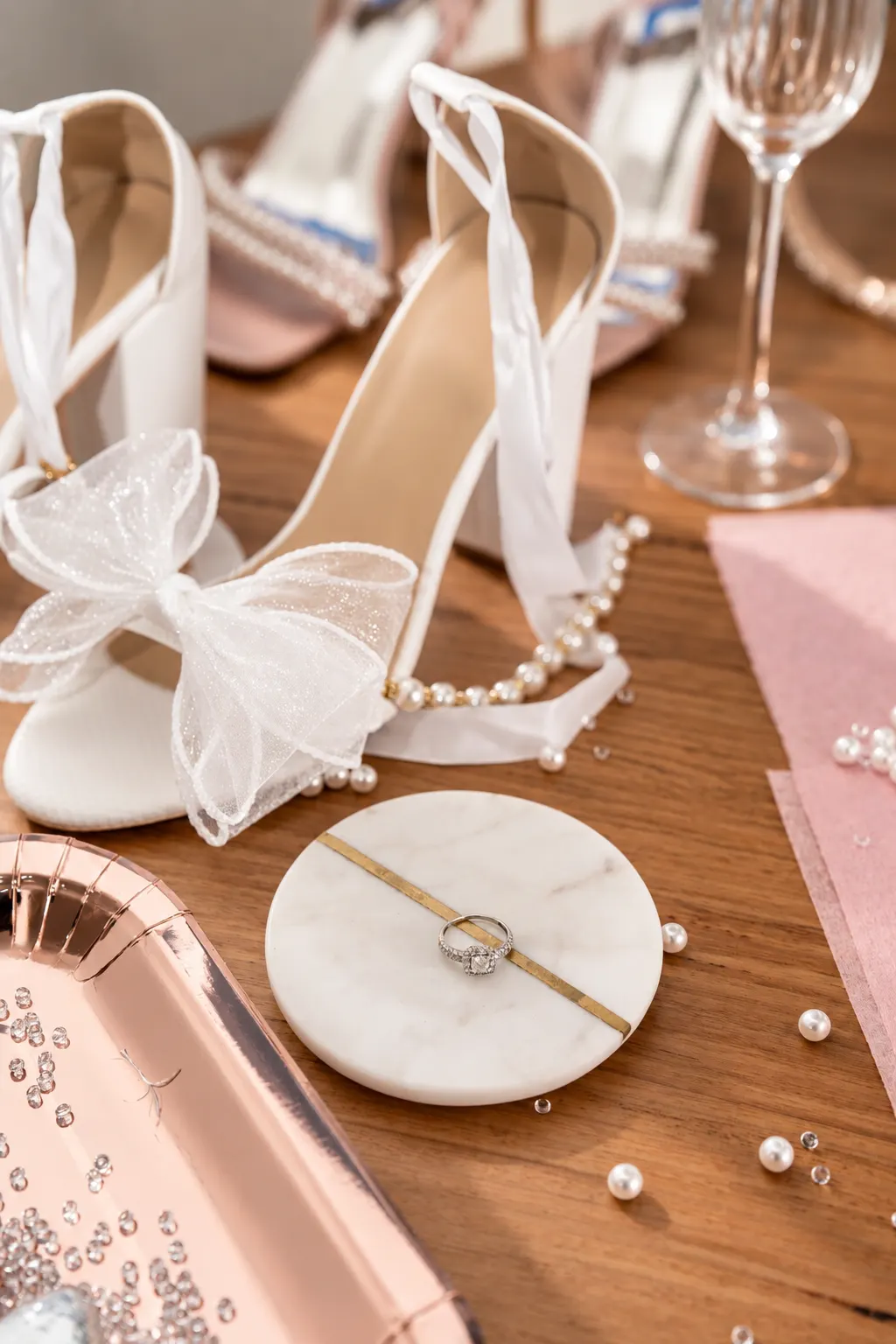 White bridal heels with bow details, a ring and pearl accessories arranged on a wooden table.