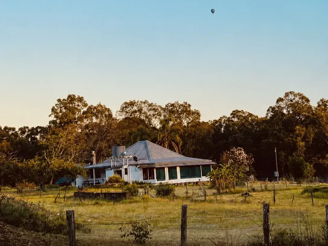 The Adeline Estate farmhouse below the treeline with a hot air balloon drifting overhead at sunrise