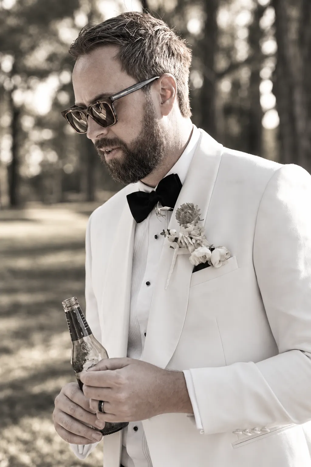 Groom in a white tuxedo, black bow tie and sunglasses standing beneath the trees.