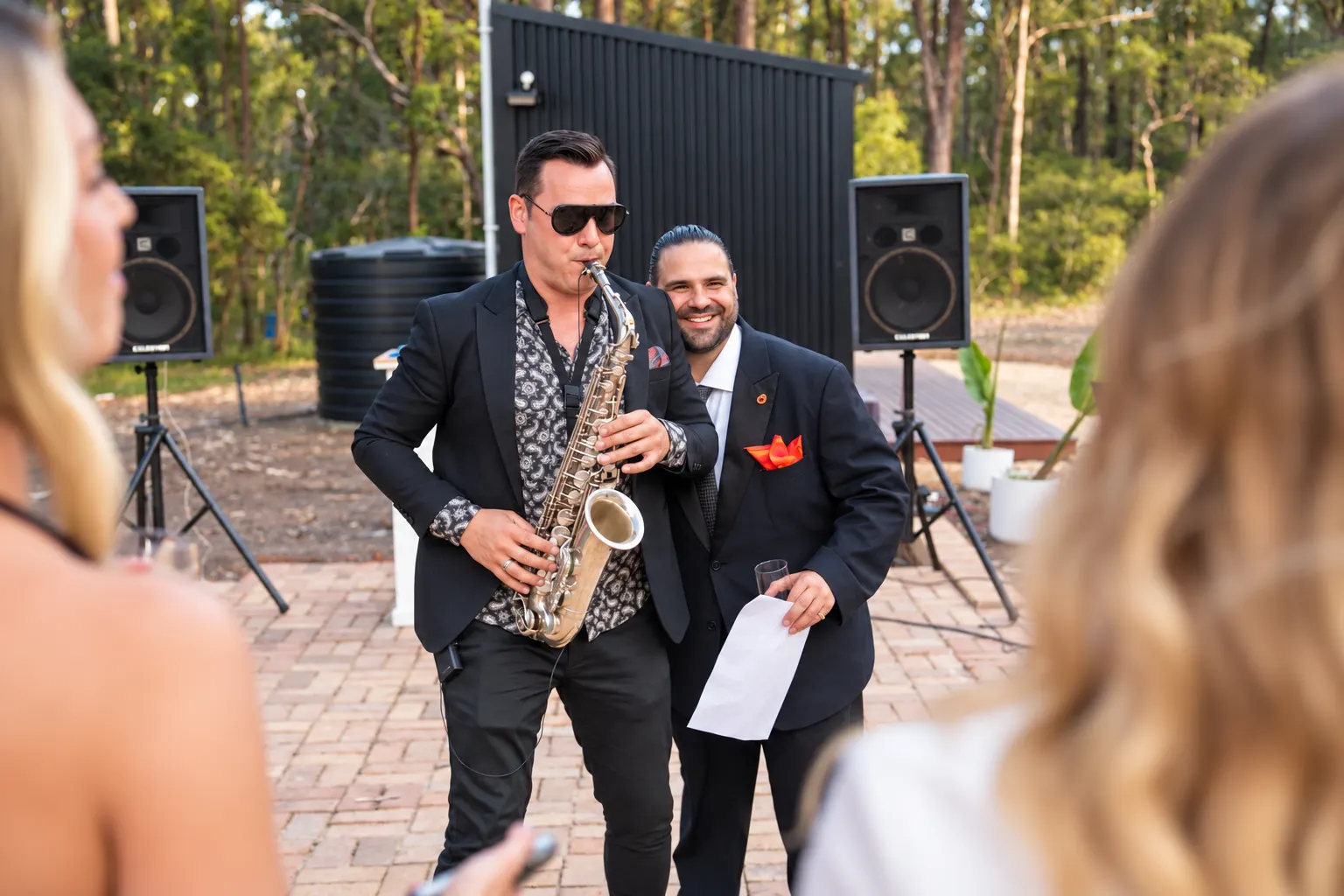 Saxophonist performing among wedding guests on a brick-paved outdoor area.