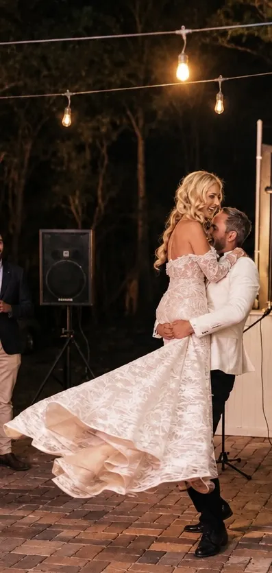 Groom lifting the bride as they dance on a brick-paved outdoor area beneath festoon lights at night.