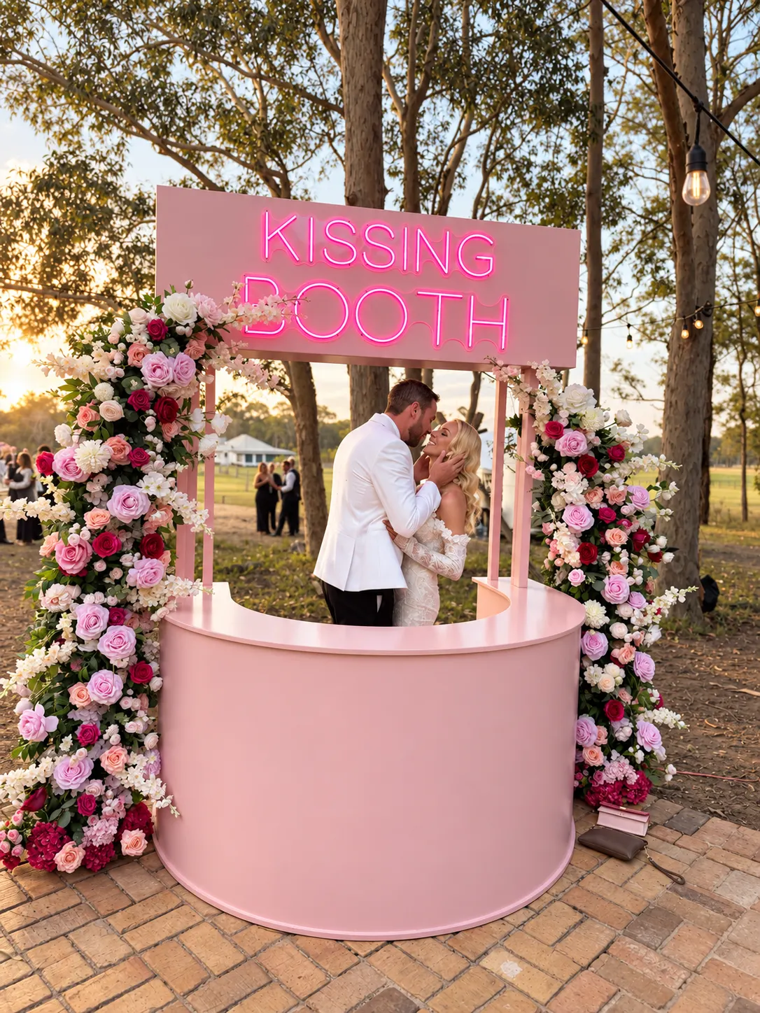 Wedding couple kissing at a pink kissing booth framed by flowers and trees.
