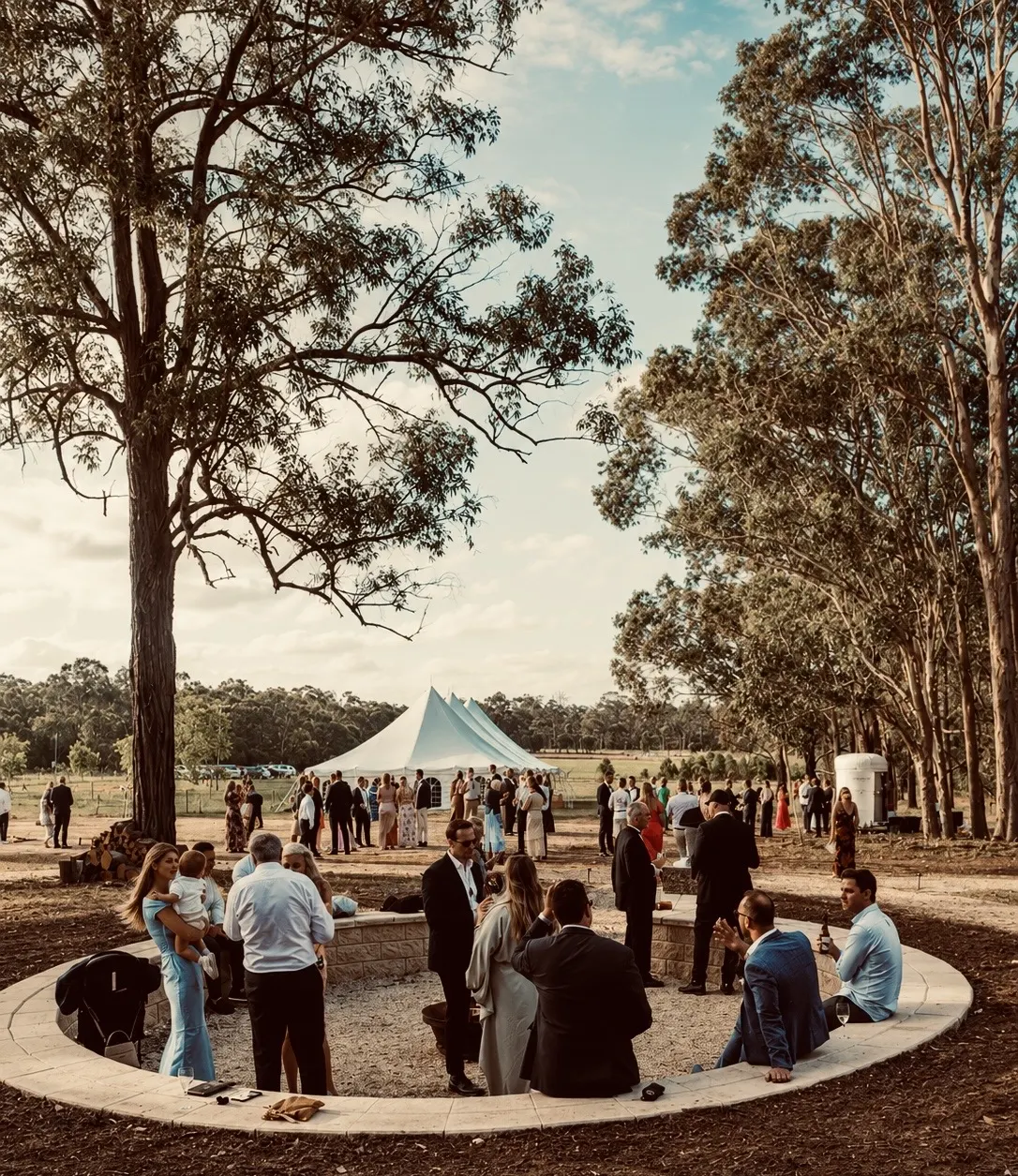 Wedding guests gathered around the circular fire-pit seating with the white marquee beyond at Adeline Estate
