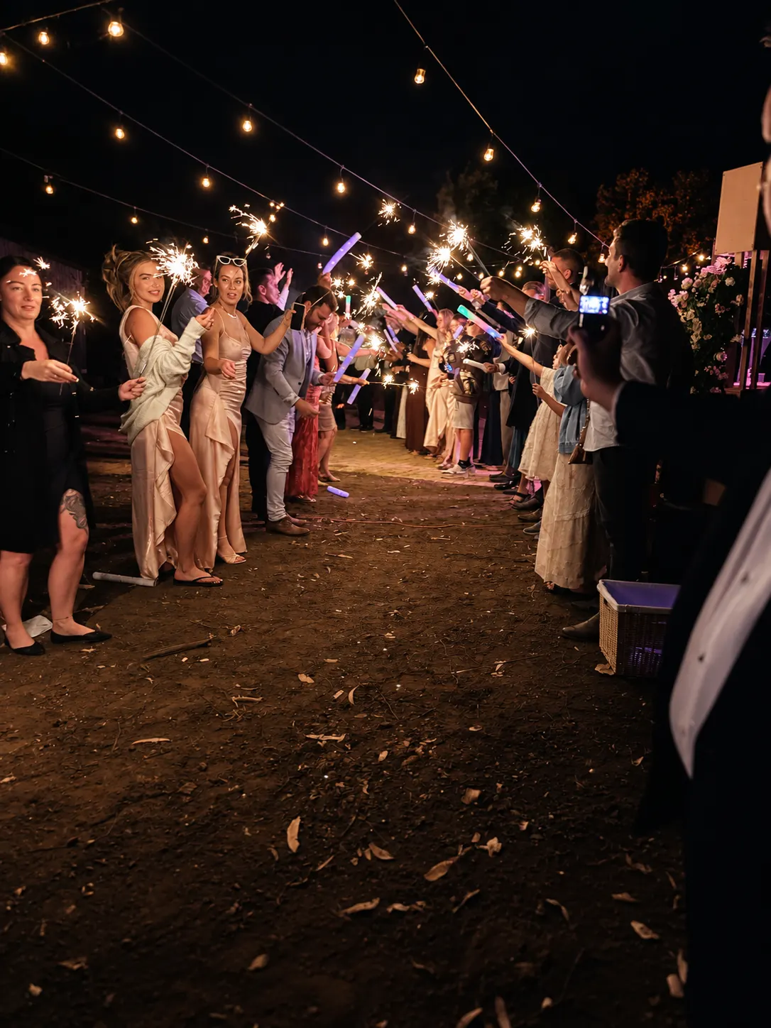 Wedding guests standing in two rows with sparklers and glow sticks beneath outdoor festoon lights at night.